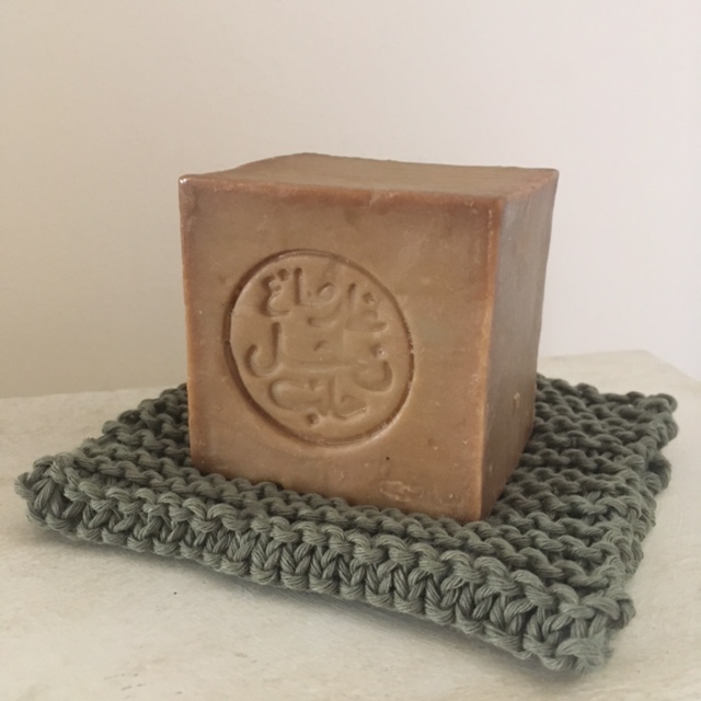 A cube of clay coloured soap, stamped with a logo featuring Arabic script, it sits on a khaki, knitted soap dish.
