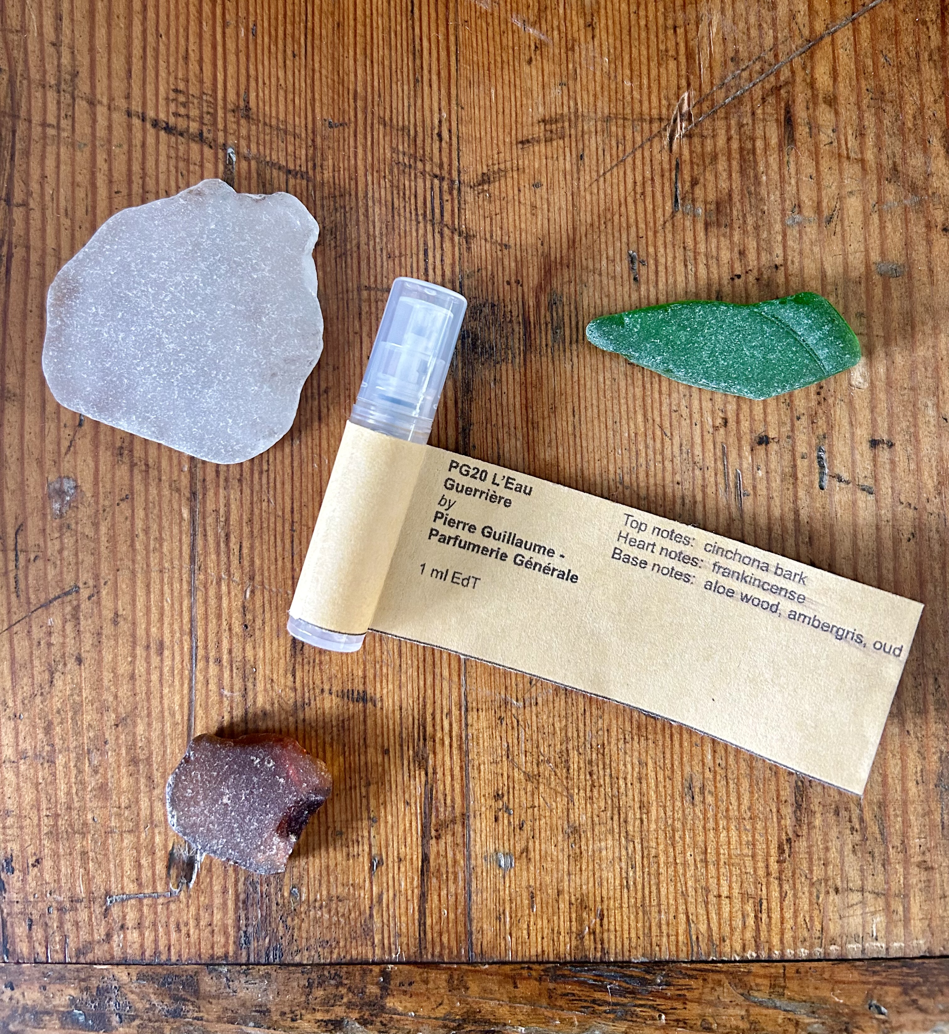 Photograph: Perfume sample vial on mid-tone wood background accompanied by three pieces of sea glass.