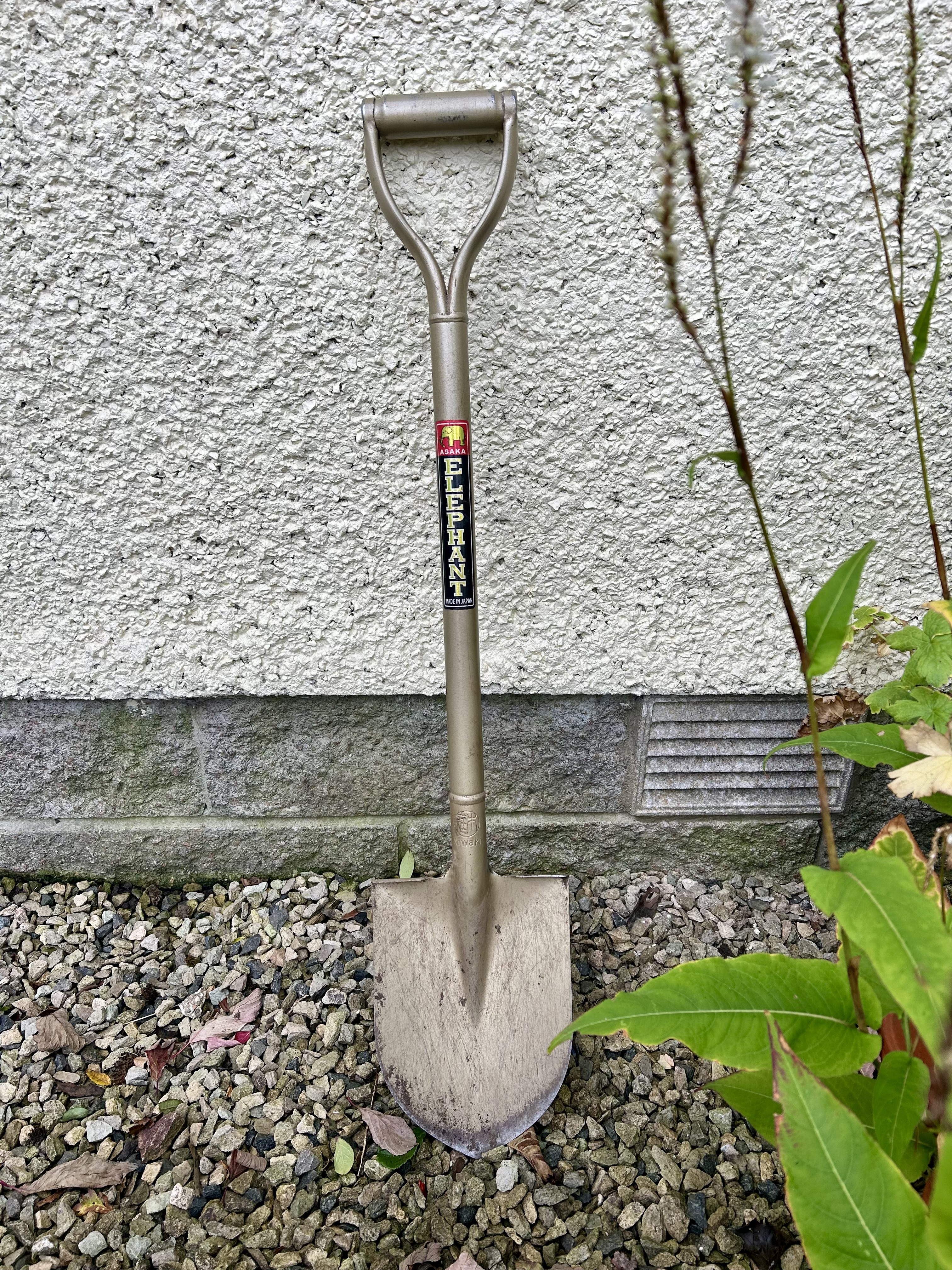 A bronzey spade leans against a cream wall.