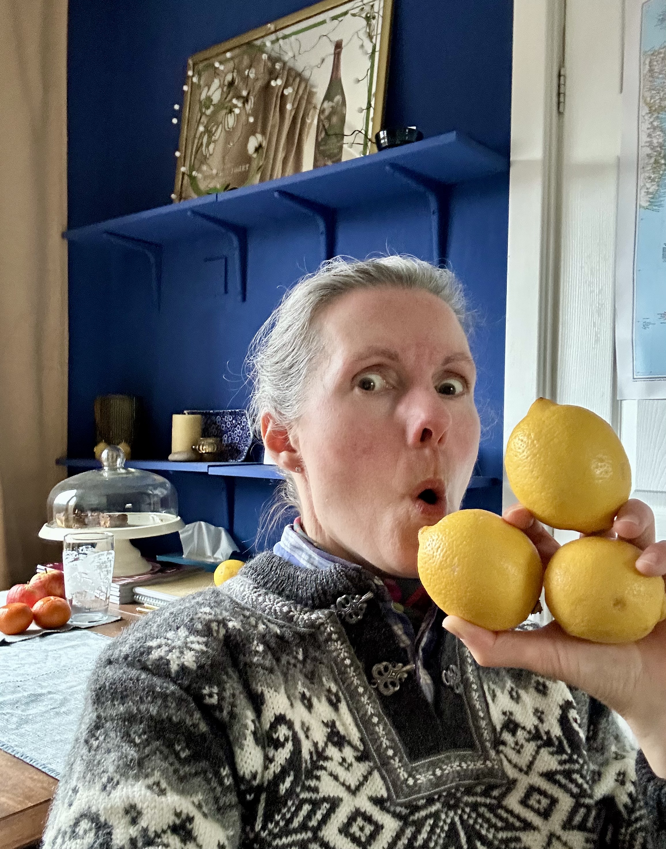 photograph of Rowan Ambrose holding a trio of bright yellow lemons next to their face, pulling a "sour" expression