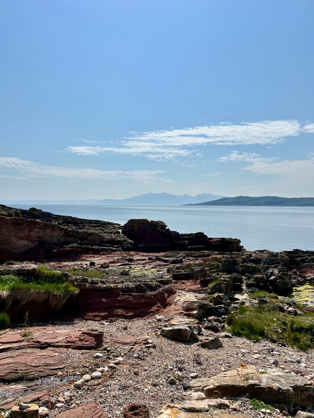 A tranquil Scottish beach, with calm, pale blue sea, hazy island mountains in the distance and vivid blue sky.