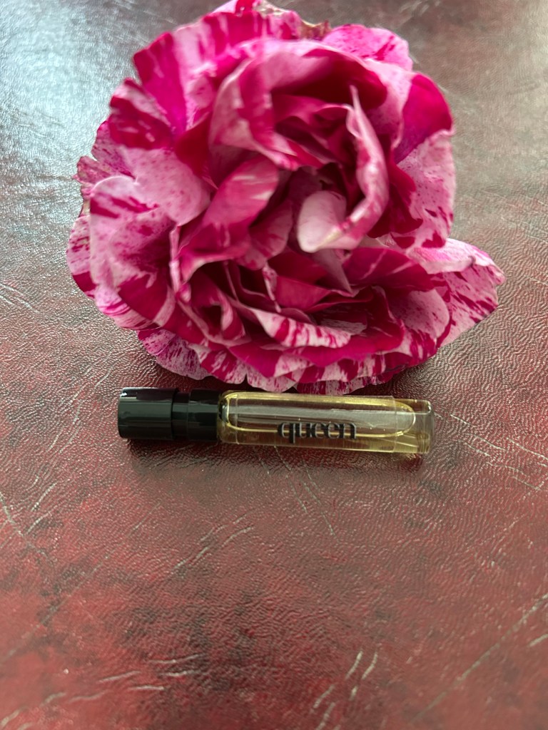 A glass vial of Queen perfume by Eau de Boujee is lying next to a raspberry pink splashed rose on a dark red leather topped desk.