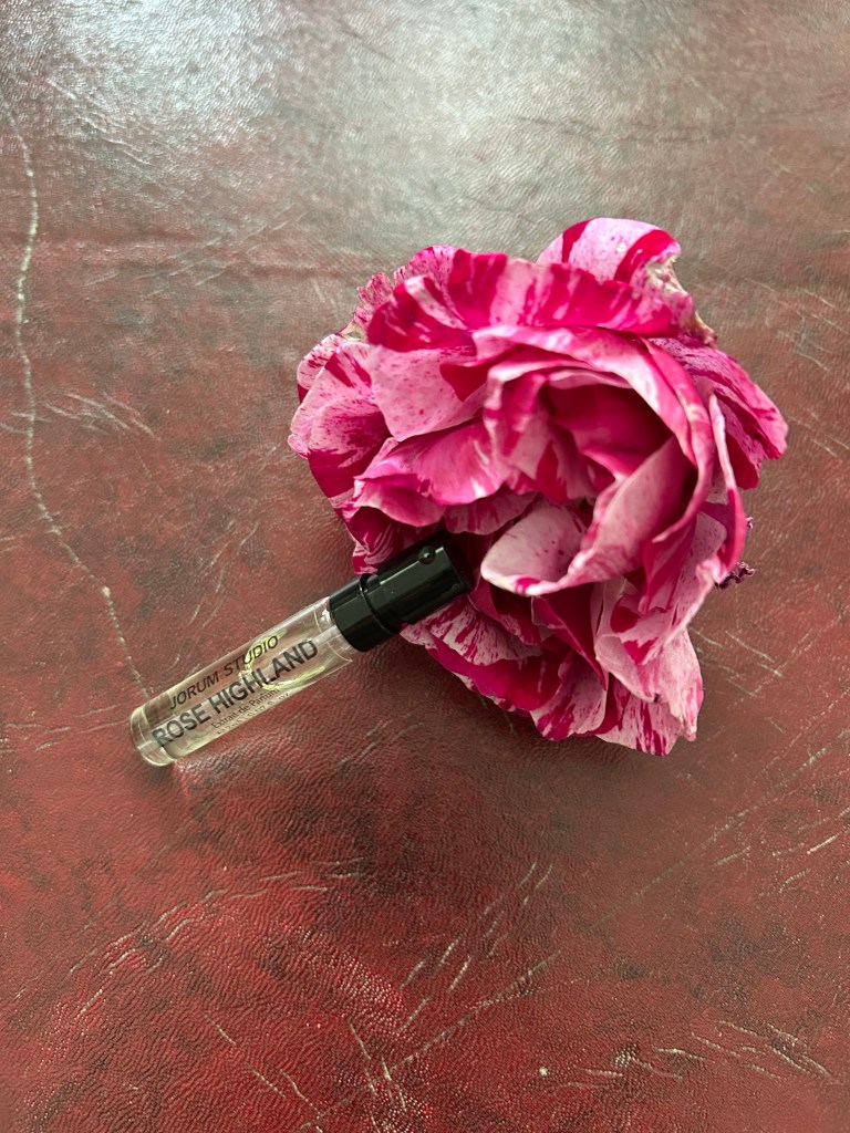 A glass vial of Jorum Studio Rose Highland perfume is propped on a raspberry pink and soft pink rose on a dark red leather topped desk.