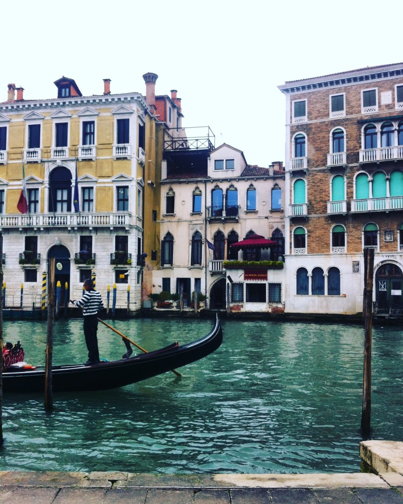 A gondola on a calm blue green canal in Venice. There are very old buildings behind, and the sky is very pale grey.