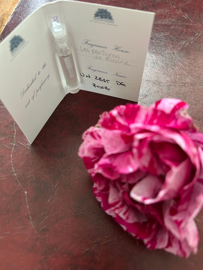 A glass vial of Un Zest de Rose perfume is attached to a small card. There's an out of focus raspberry pink and soft pink streaked rose on the dark red leather topped desk.