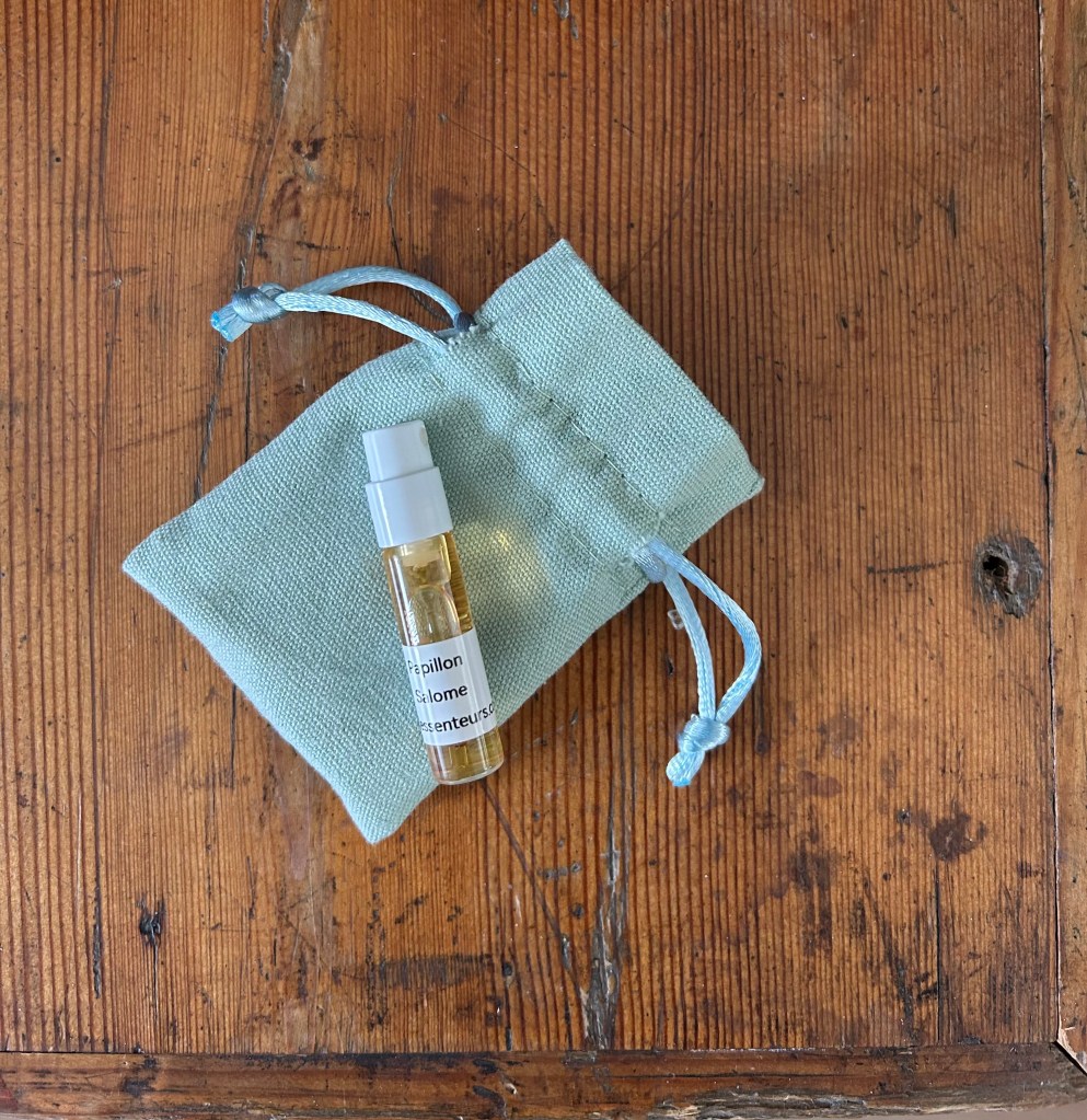 A glass vial of Salome perfume by Papillon is lying on a duck egg blue linen pouch, which is on an antique pine trunk.