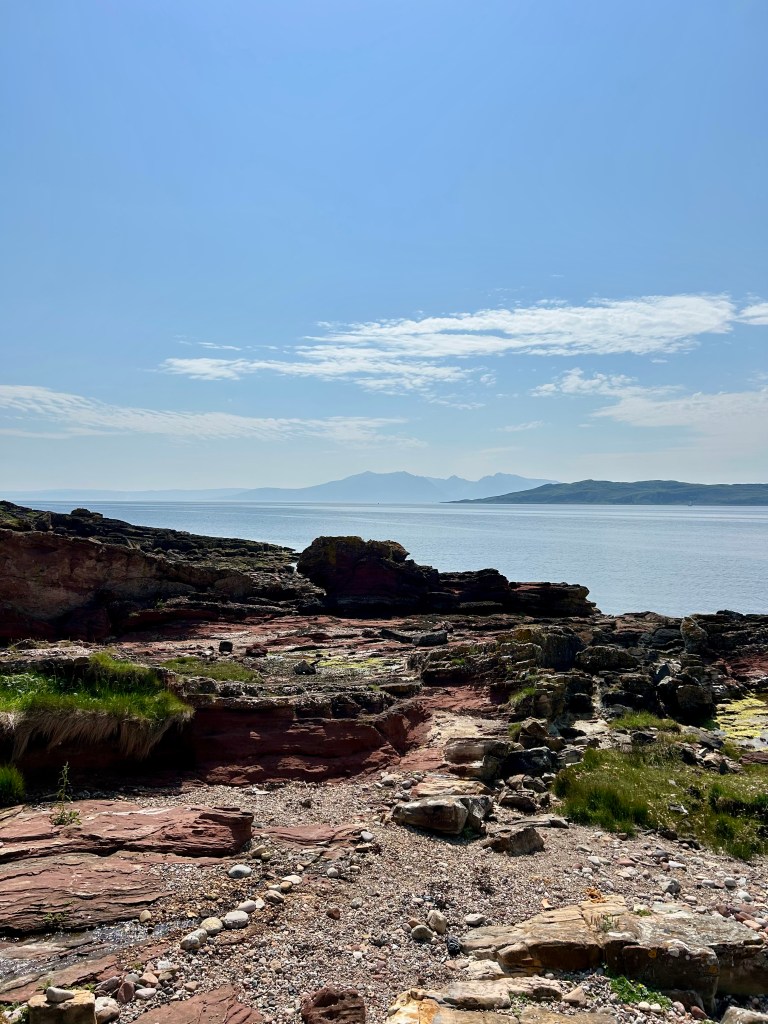 Clear blue skies over the hazy and distant mountain peaks on the Isle of Arran as seen from a red, rocky beach on the Isle of Cumbrae. There is shingle on the beach and odd tufts of wiry grass.