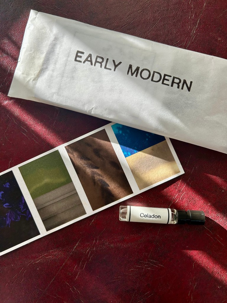 A glass vial of Celadon perfume from Scottish fragrance house Early Modern is lying on a dark red leather topped desk next to a card with four different coloured abstract images.