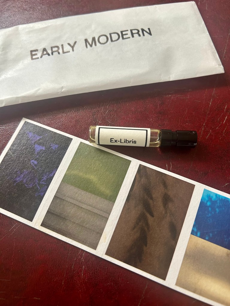 A glass vial pf Ex Libris perfume by Early Modern is lying on a dark red leather topped desk next to a waxed paper sleeve with Esrly Modern branding on it and a card with four different coloured abstract images.