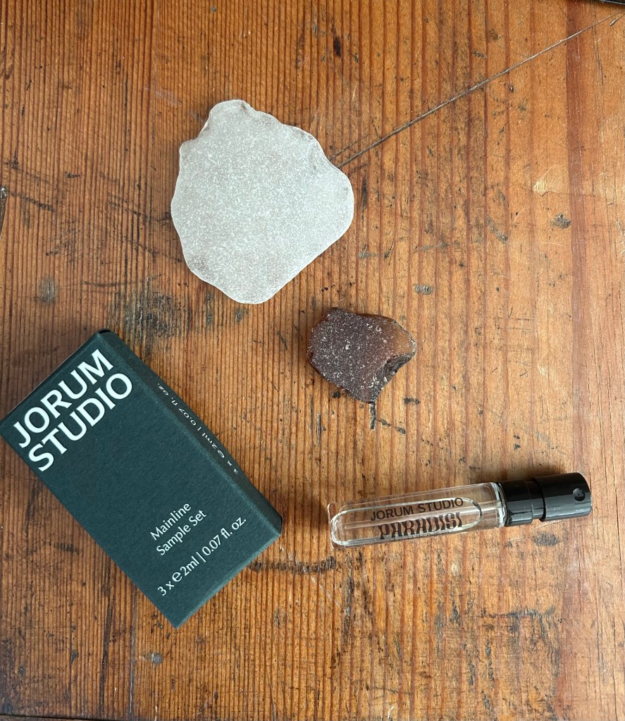 A glass vial of Paradisi perfume from Scottish fragrance house Jorum Studio is on an antique pink trunk. There are brown and clear pieces of sea glass and a dark green Jorum Studio box.