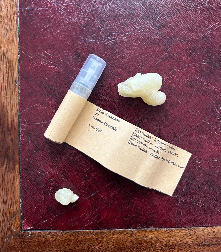 A small plastic vial of perfume from Naomi Goodsir is lying on a dark red leather topped table, surrounded by ice green chunks of genuine frankincense.