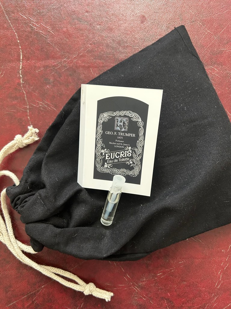 A small glass vial of Eucris by Geo. Trumper is lying on a black fabric bag with the brand packaging next to it. Everything is on a dark red leather topped desk.