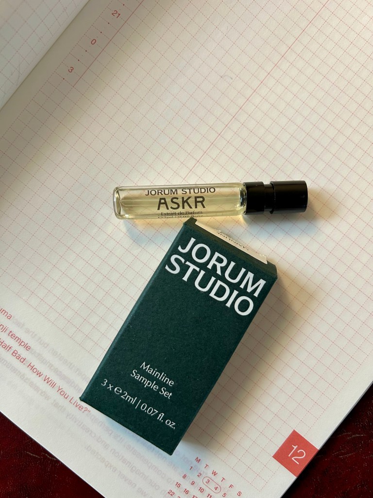 A glass vial of Askr perfume by Jorum Studio is on a grid marked notebook next to a dark green box with Jorum Studio branding.