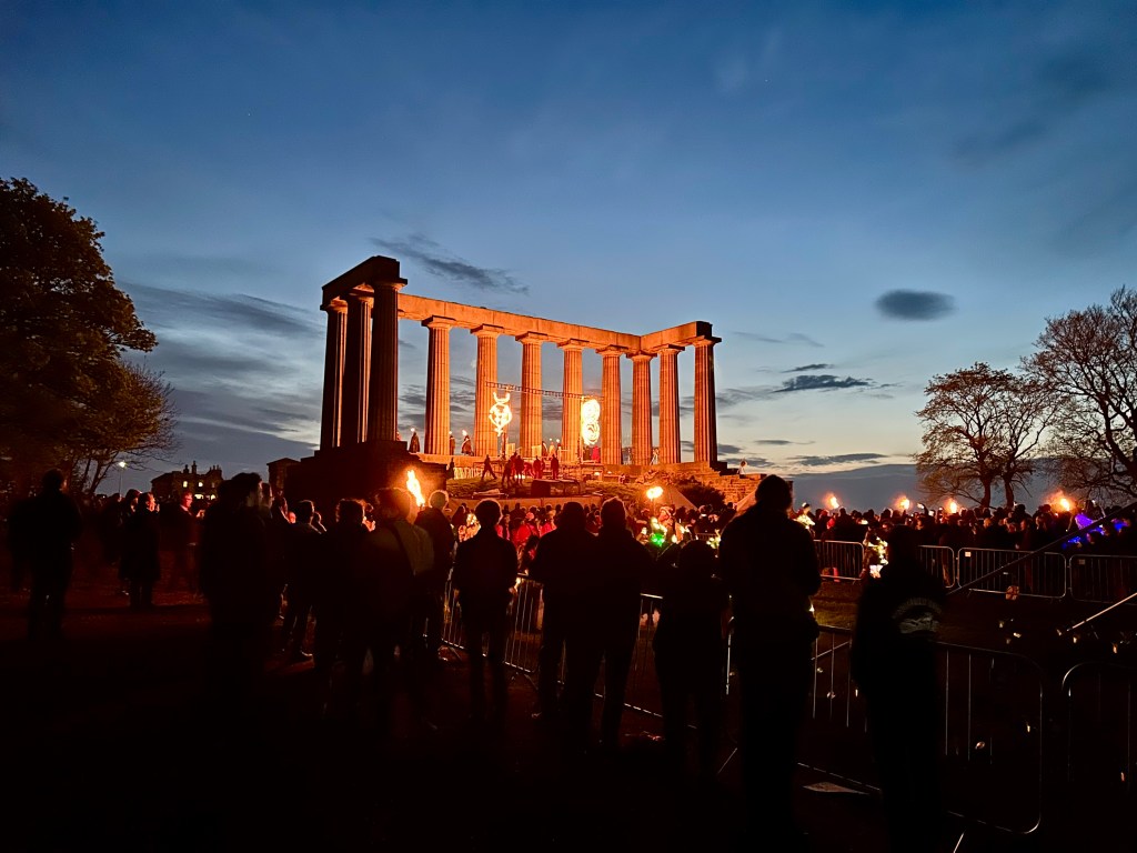 The Beltane Fire Festival on Calton Hill in Edinburgh, shows an ancient monument lit with fire and fireworks against a darkening sky. There are lots of people gathered to watch the performers.