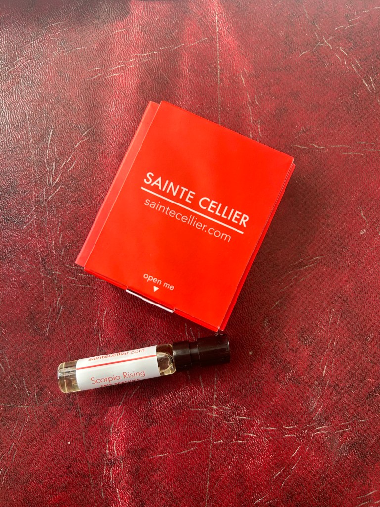 A glass vial of Scorpio Rising by ERIS Parfums is next to a bright red box with Sainte Cellier branding. They're both on a dark red leather topped desk.