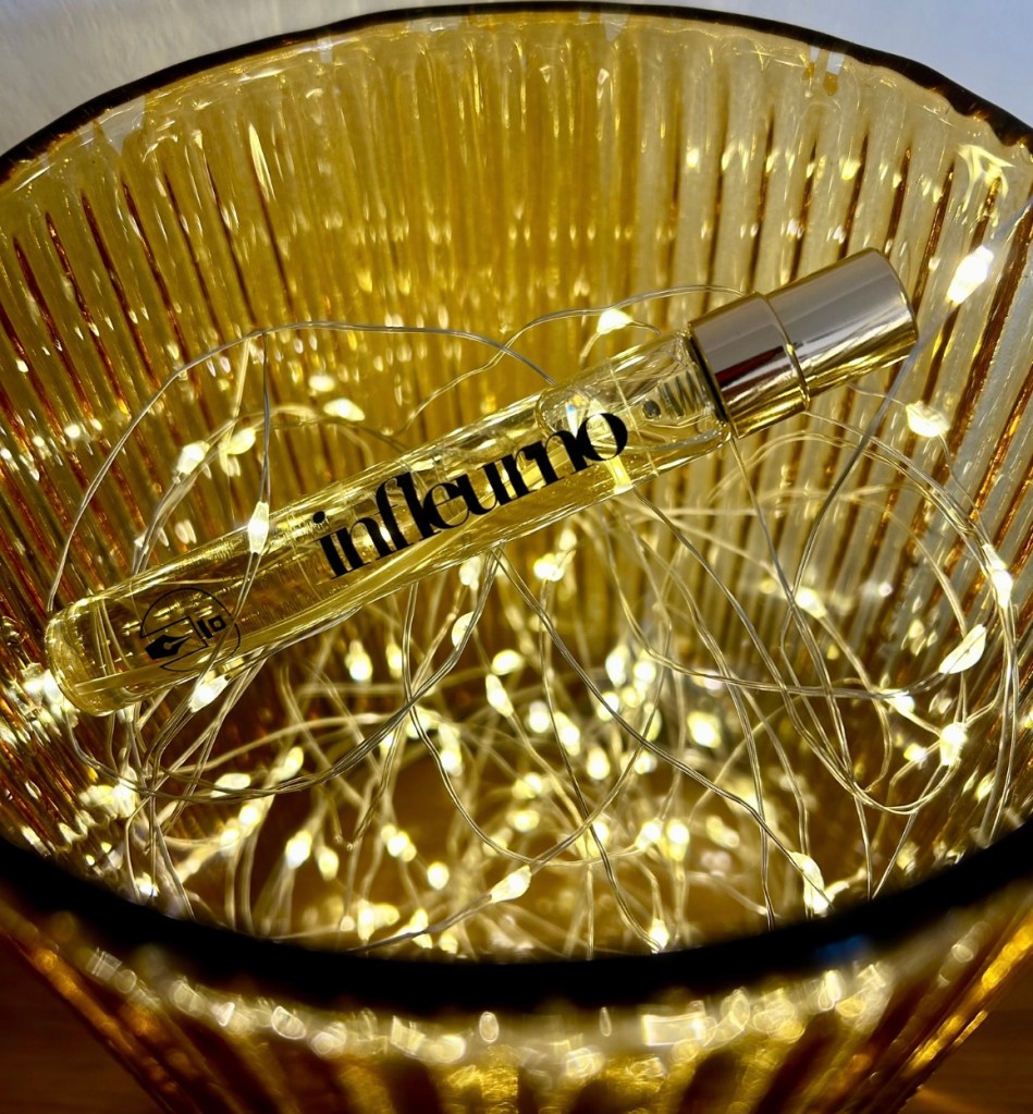 A travel size glass bottle of Infleurno perfume by Eau de Boujee is in an amber candle holder filled with tiny golden lights. This fragrance has a prominent passionfruit note.
