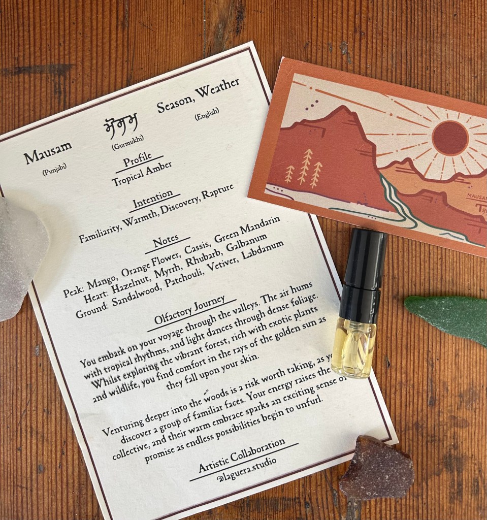 A small glass bottle of Mausam perfume by Kushbu is surrounded by Kushbu cards with branding images of sun and mountains, and pieces of sea glass. Everything is on an antique pine trunk.