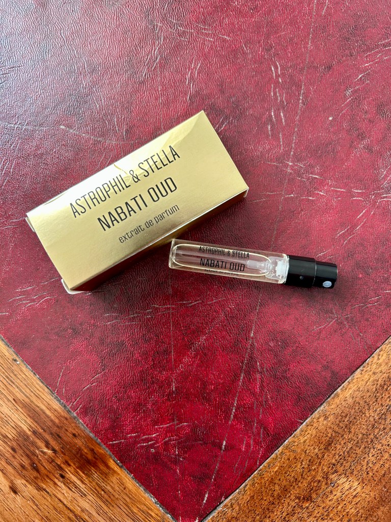 A small glass vial of Nabati Oud perfume by Astrophil and Stella is on a dark red leather topped desk next to a gold box with Astrophil and Stella branding on.