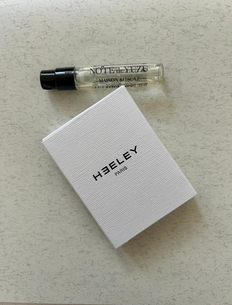 A small glass vial of Note de Yuzu perfume by Heeley Parfums is on a marbled grey notebook next to a white box with black Heeley branding on.
