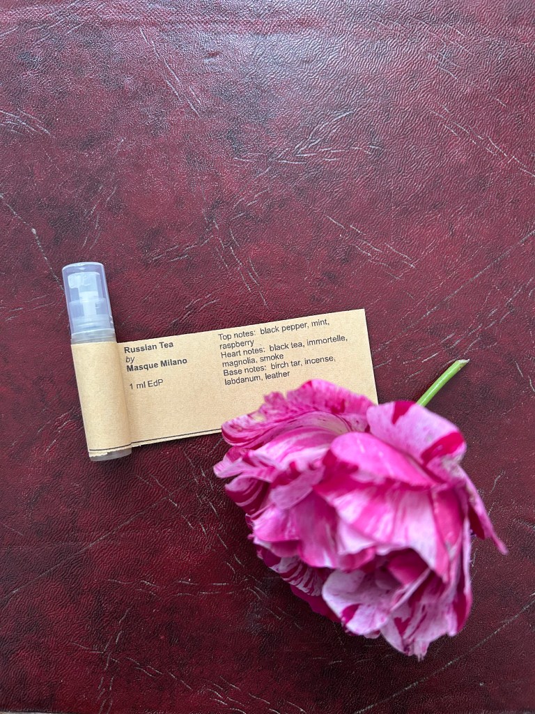 A small plastic vial of Russian Tea perfume by Masque Milano is on dark red leather topped desk. There's a raspberry pink rose next to it as the perfume has a fabulously fruity raspberry note.