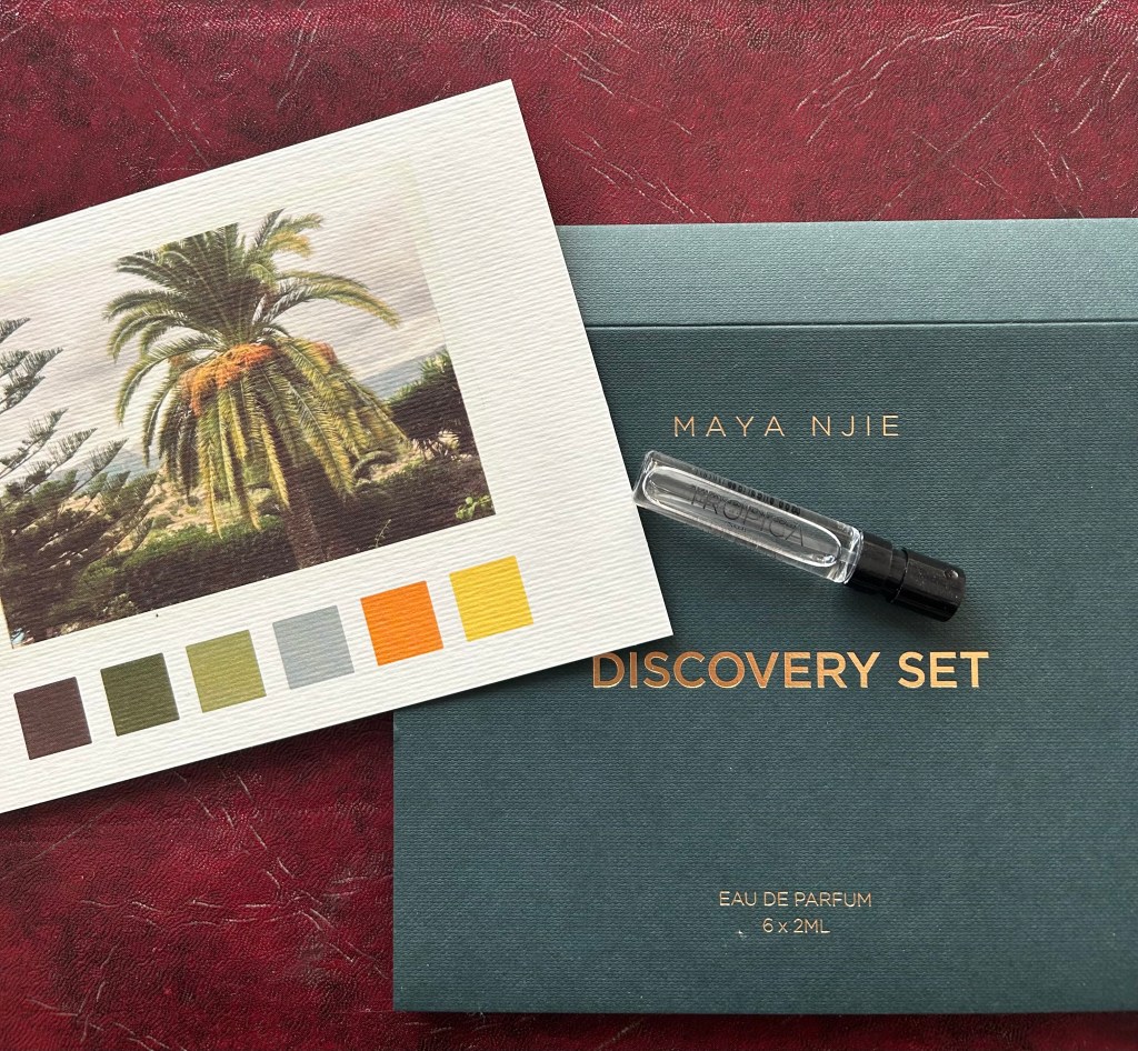 A small glass vial of Tropica perfume by Maya Njie is on a dark green box with gold Maya Njie branding. There's a postcard with a picture of a graceful palm tree and squares on colour next to it.