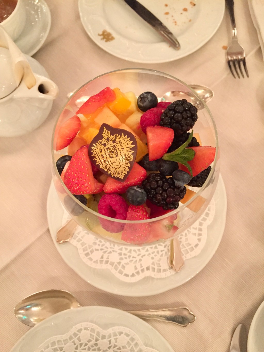 A fine glass dish with small and colourful pieces of fresh fruit sits on a white tablecloth surrounded with white china and silver cutlery.