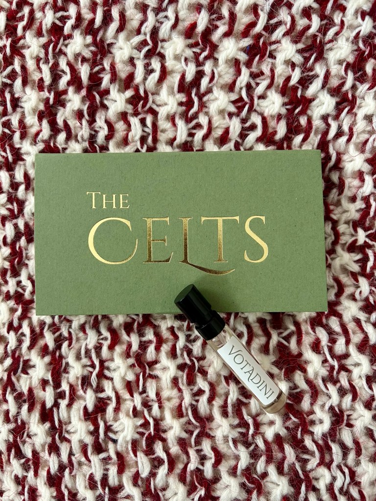 A small glass vial of Votadini perfume by Wales Perfumery is next to a green box with Wales Perfumery branding in gold. This fragrance has a bold dried fruit note. Everything is on a dark red and cream knitted cushion.