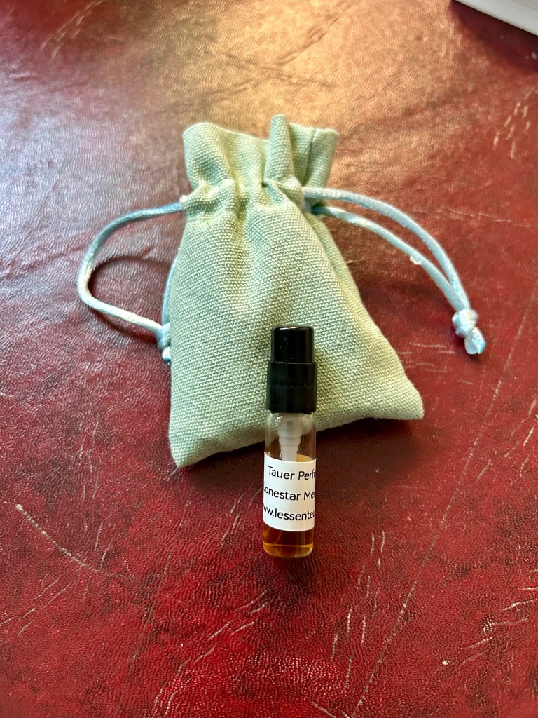 A glass vial of Lonestar Memories by Any Tauer is resting on a sage green linen pouch on a dark red leather topped desk.