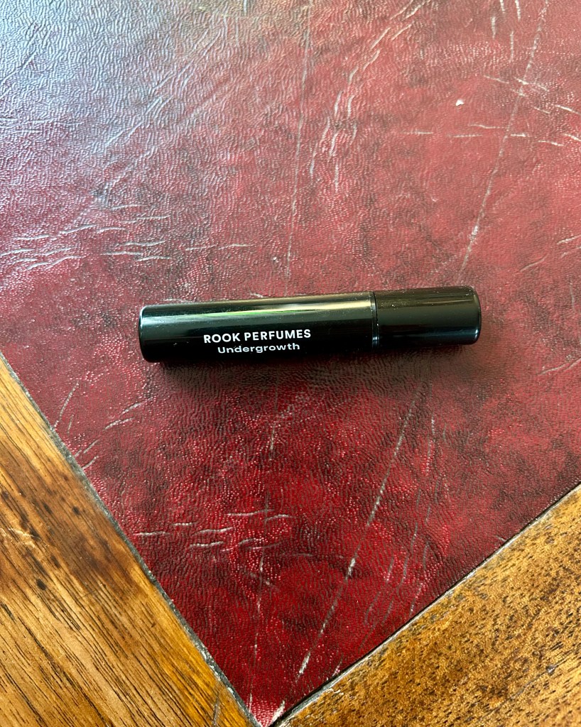 A black plastic vial of Undergrowth perfume by Rook sits on a dark red leather topped desk.