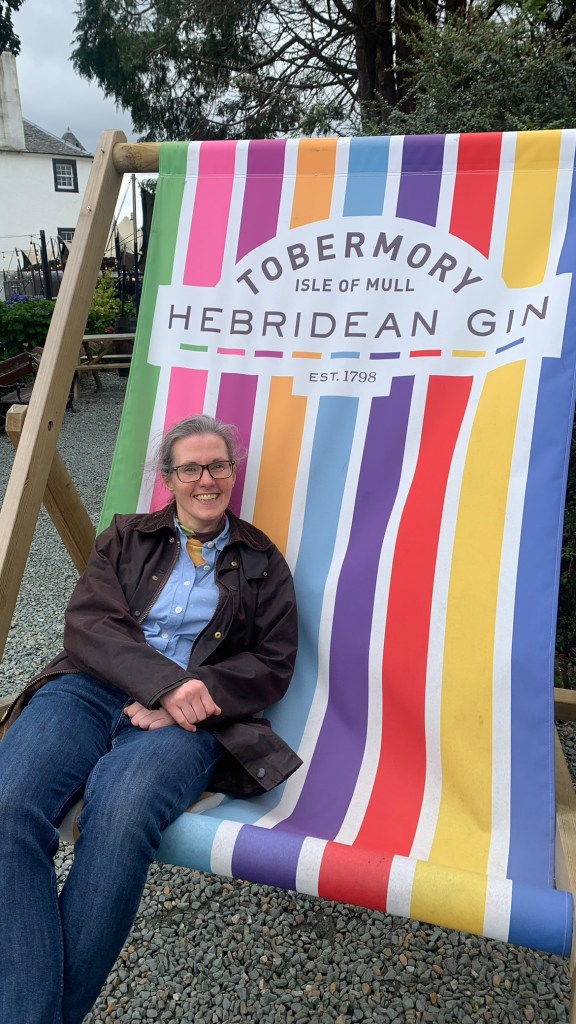 Rowan Ambrose is reclining on a gigantic, striped deckchair with Tobermory Gin branding.