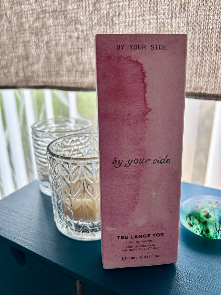 A marbled pink cardboard box of By Your Side perfume is sitting on a teal wooden box by textured glass candle holders.