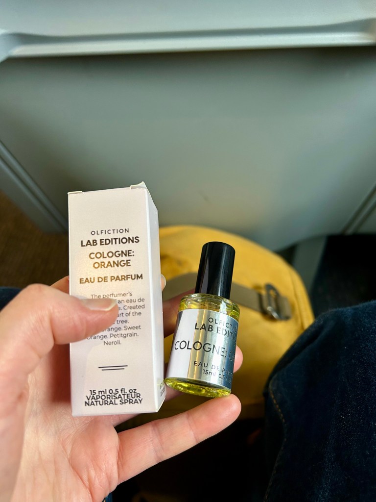A white person's hand holds a small glass bottle of Cologne: Orange perfume. There's a grey train seat back and a yellow backpack behind. 