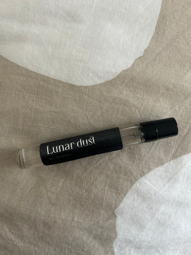 A small glass bottle of Lunar Dust perfume is lying on stone and cream coloured fabric.