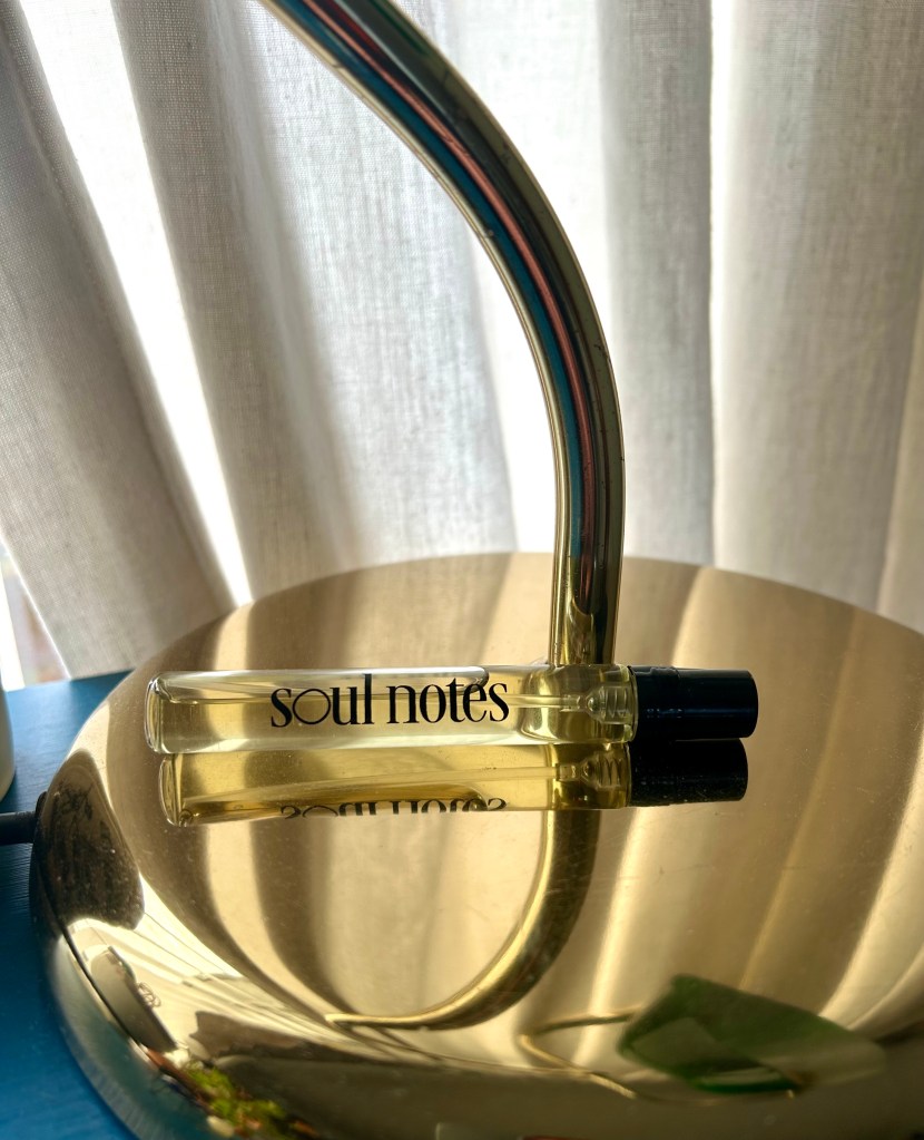 A small glass bottle of perfume is lying on a brass lamp stand with cream linen curtains behind.
