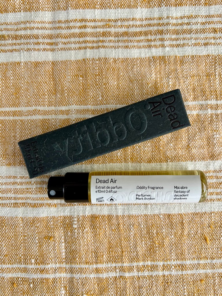 A travel size glass bottle of Oddity perfume is by its dark grey box on a gold and white linen cloth.