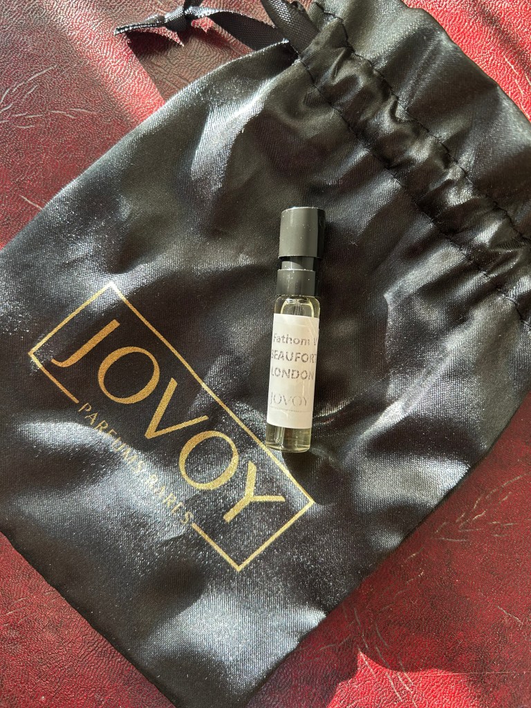 A small glass vial of perfume on a black satin Jovoy bag on a dark red leather topped desk.