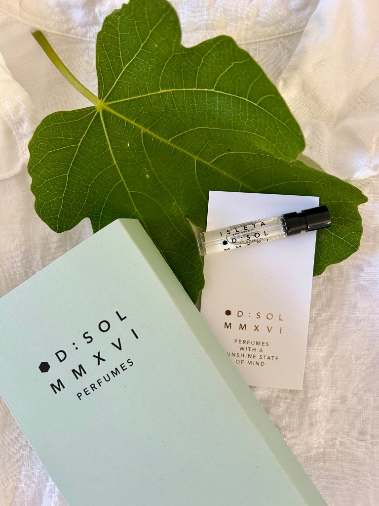 A small glass vial of Isleta perfume is next to its pale blue packaging on a fresh fig leaf. Everything is lying on a crisp white linen shirt.