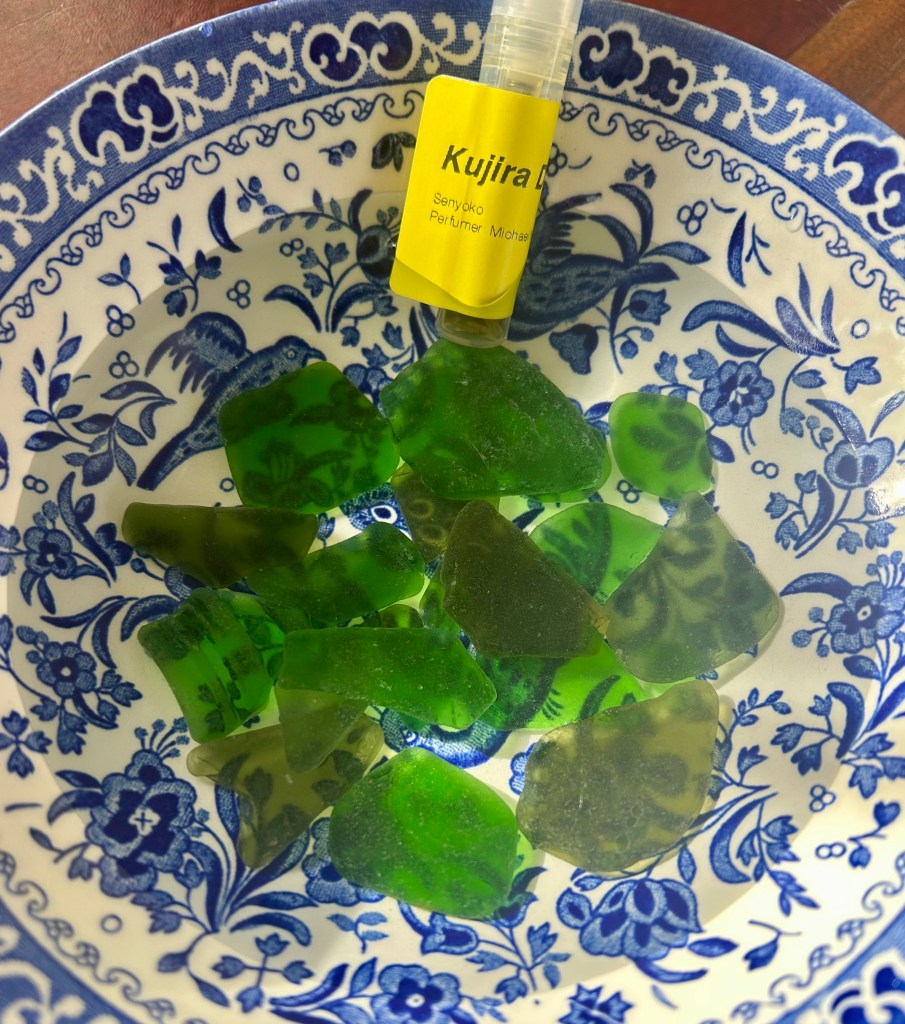 A small vial of perfume sits in a blue and white bowl filled with water and bright green sea glass.