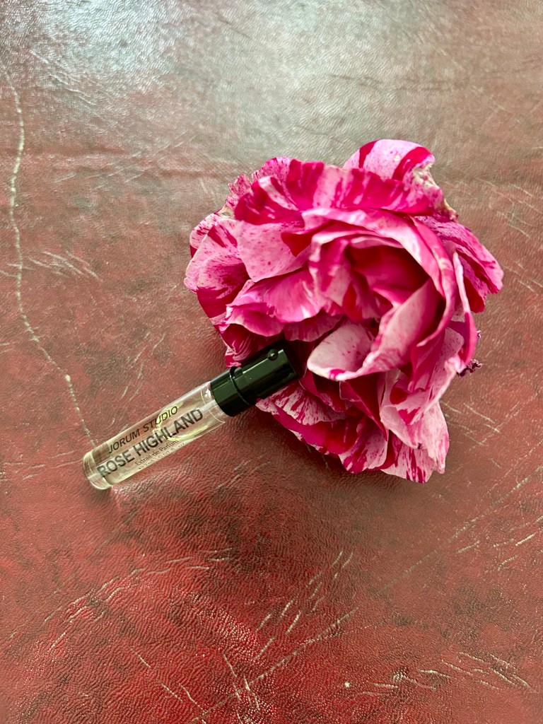 A raspberry rippled rose has a glass vial of perfume leaning on it. They're both on a dark red leather topped desk.
